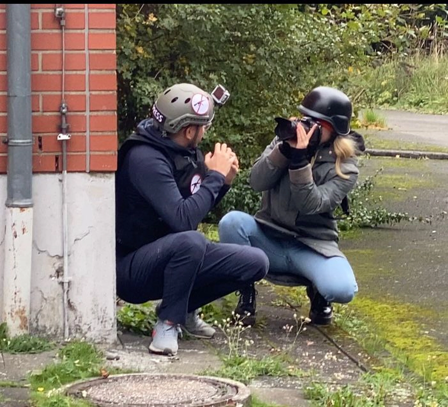 Krisenwerk GmbH - Crisis management training - A man in a white helmet with a camera attached, wearing a navy hoodie and jeans, crouching and gesturing with his hands toward a woman in a black helmet and gray jacket who is photographing.