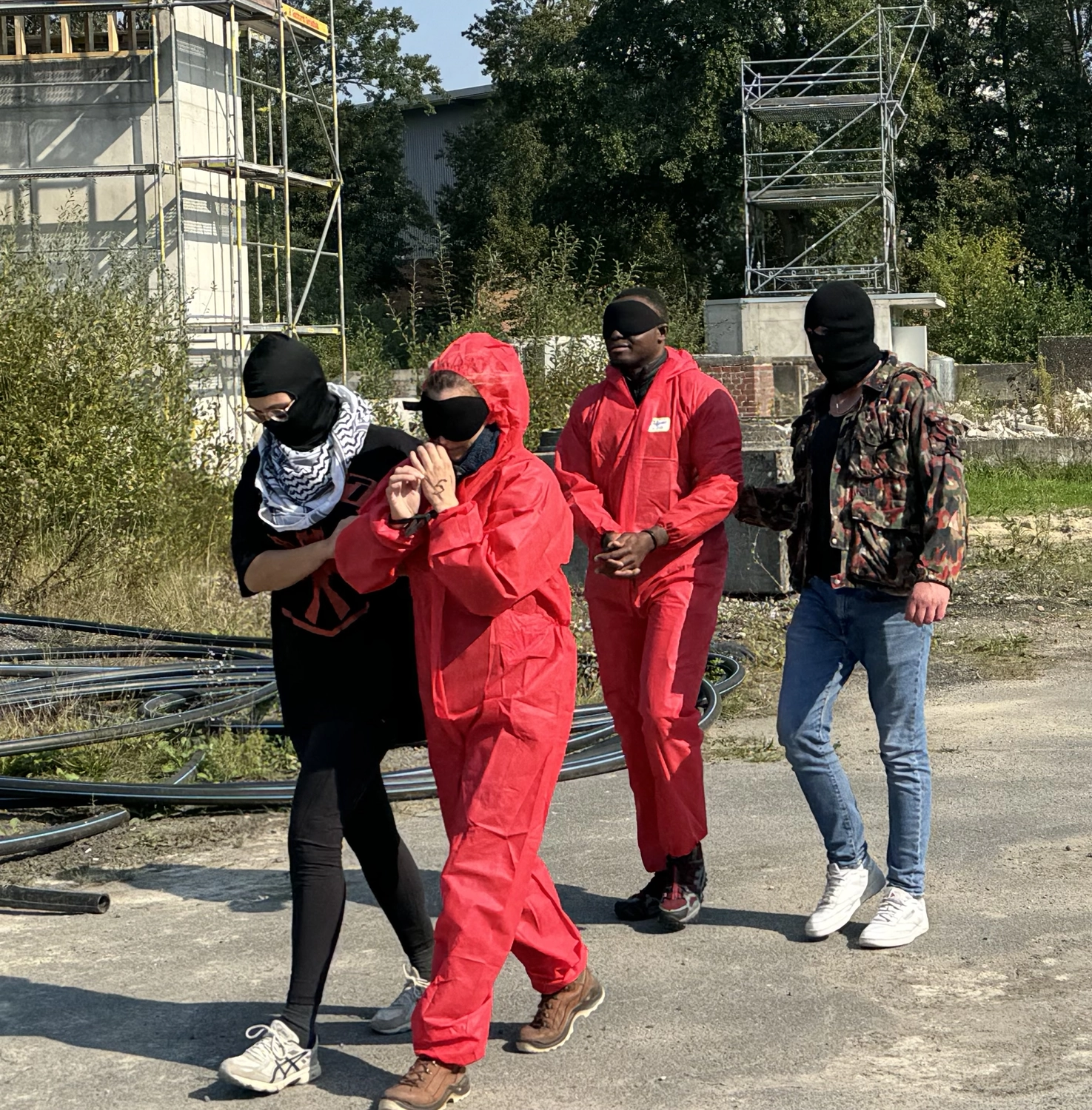 Krisenwerk GmbH - Crisis management training - Two individuals in red protective suits and black face masks stand on a road with construction scaffolding in the background. Two additional masked people in casual attire appear to be taking them anywhere.