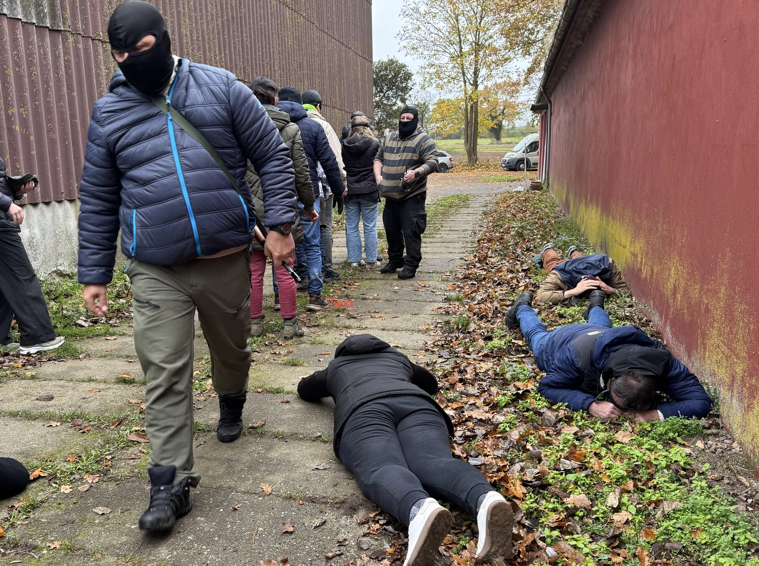 Krisenwerk GmbH - Crisis management training - A man wearing a blue jacket, gray pants, and a black mask walks on a concrete sidewalk next to three person lying face‑down on the ground among fallen leaves.