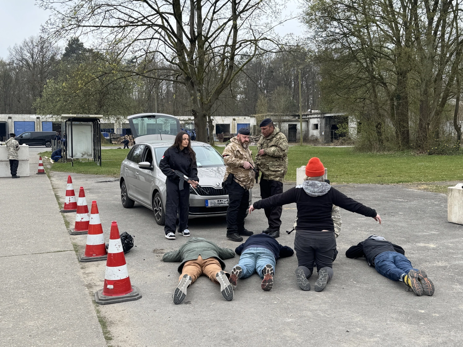 Krisenwerk GmbH - Crisis management training - An outdoor daytime setting with a woman in black clothing holding a firearm standing near a silver car parked on the road lined with red‑white traffic cones. Two individuals are lying on the pavement, and armed persons in camouflage uniforms observe the scene.