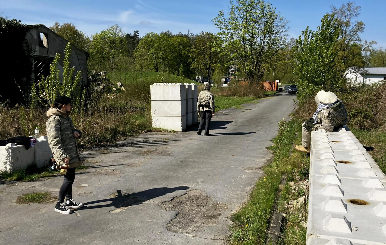 Krisenwerk GmbH - Crisis management training - A person in a gray jacket and black pants stands on road facing a building entrance, with two other individuals visible in the background near the entrance under a blue sky.